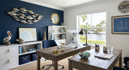 Nautical home office decor featuring a weathered wood desk, navy blue wall, and handcrafted coastal sculptures like a pelican and sea turtle. A large window overlooks a Florida waterway with palm trees and boats.