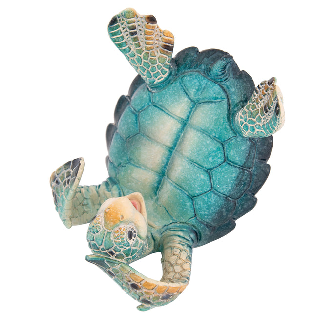 A detailed side-angle view of a textured teal and blue resin sea turtle figurine lying on its back, using its front and back flippers to securely hold a pink and black smartphone in place against a crisp white background.